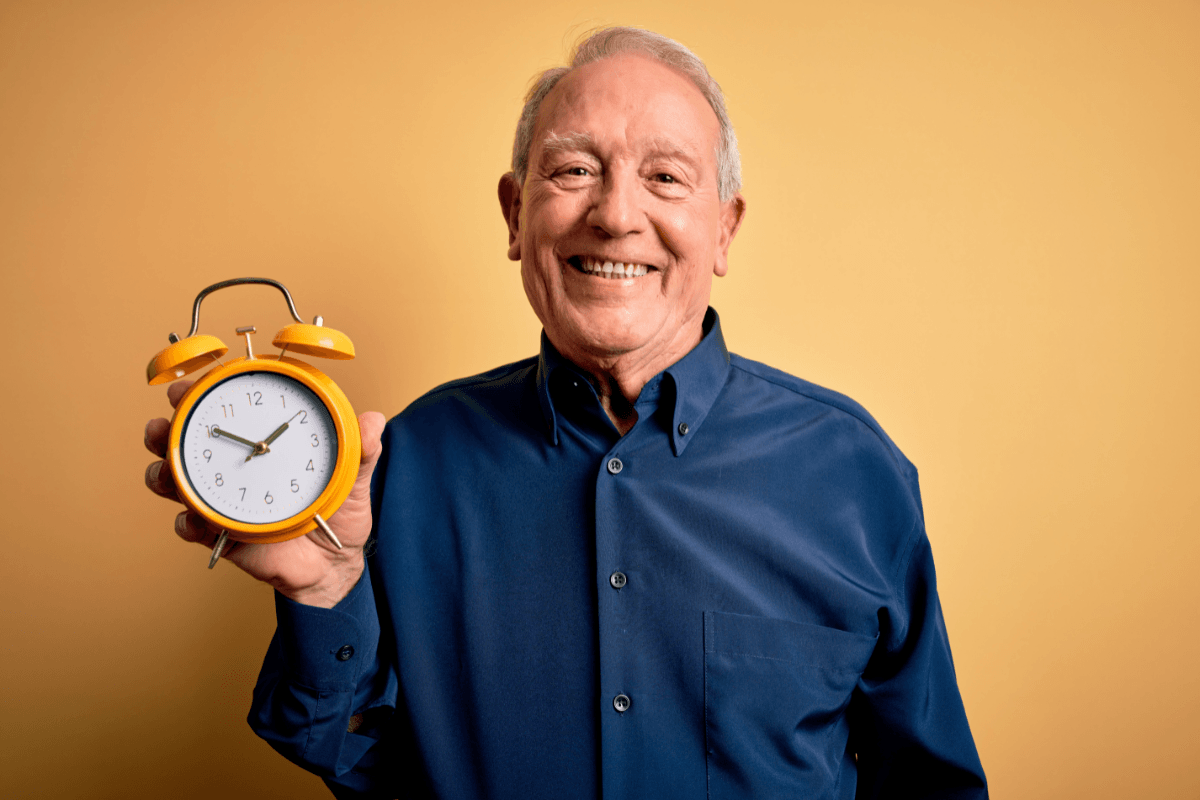 A cheerful senior man with grey hair stands confidently against a yellow background, holding a vintage alarm clock.